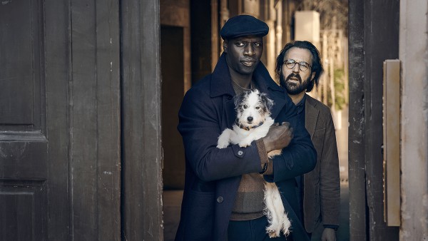 A still from the show Lupin with the titular character holding a dog while standing in a doorway.