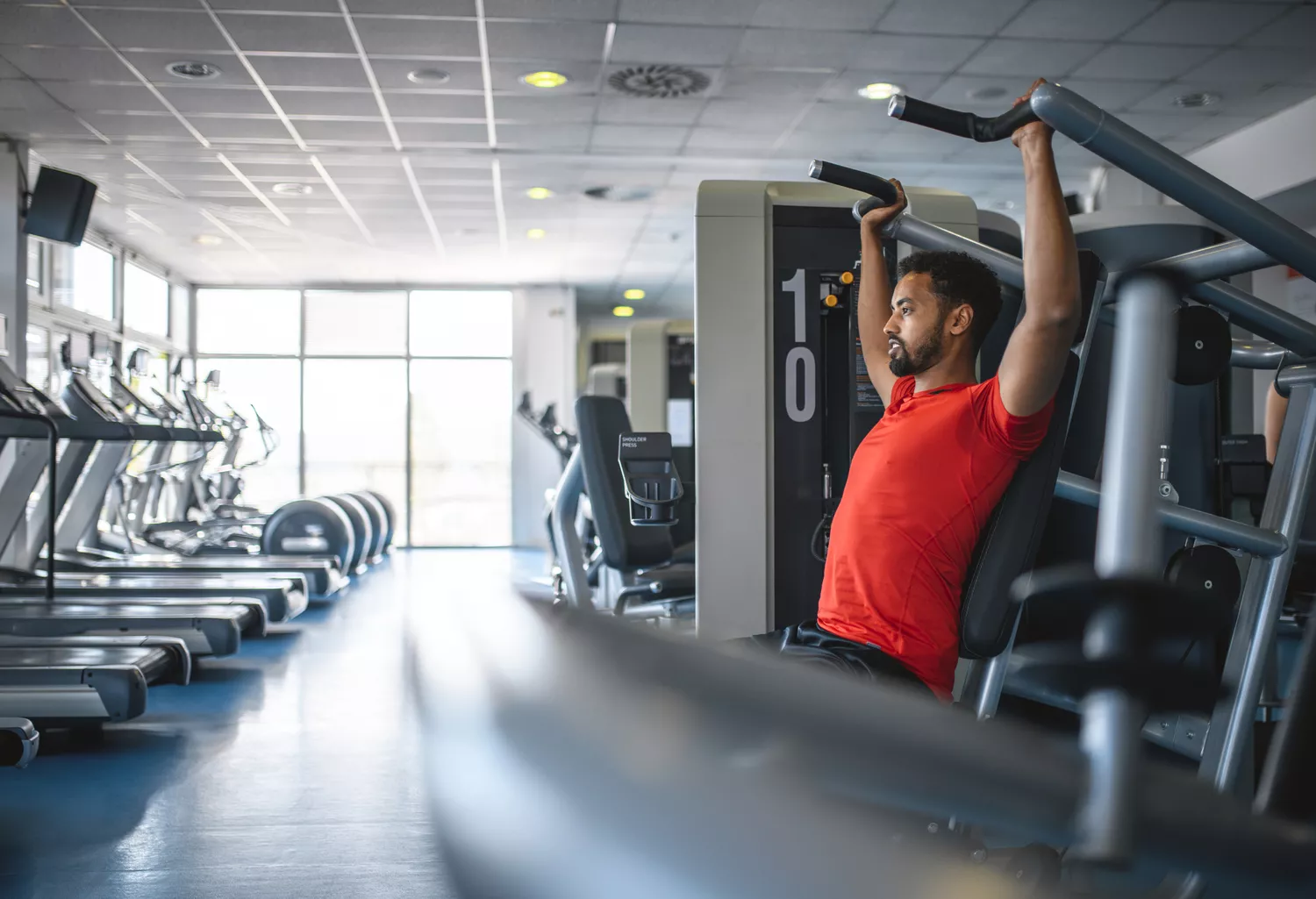 Side view of African man in early 30s working out to strengthen and tone with seated shoulder press at gym.