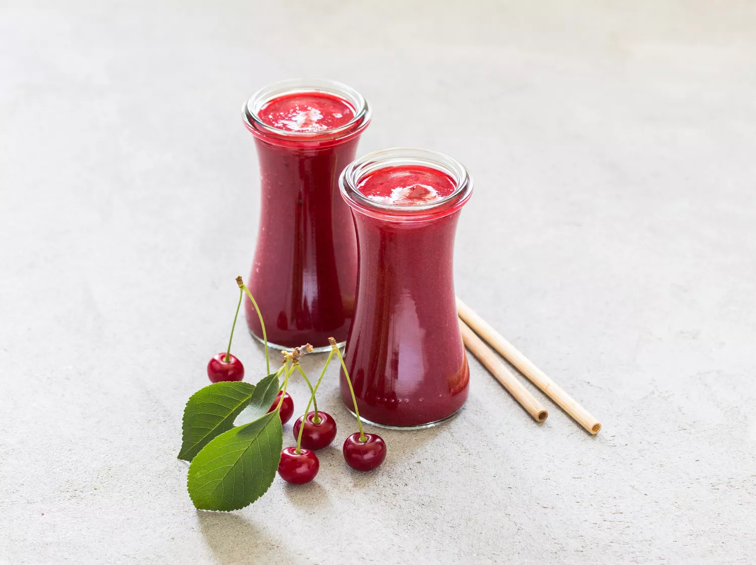 Two glasses of cherry juice with cherries and two bamboo straws on the table.