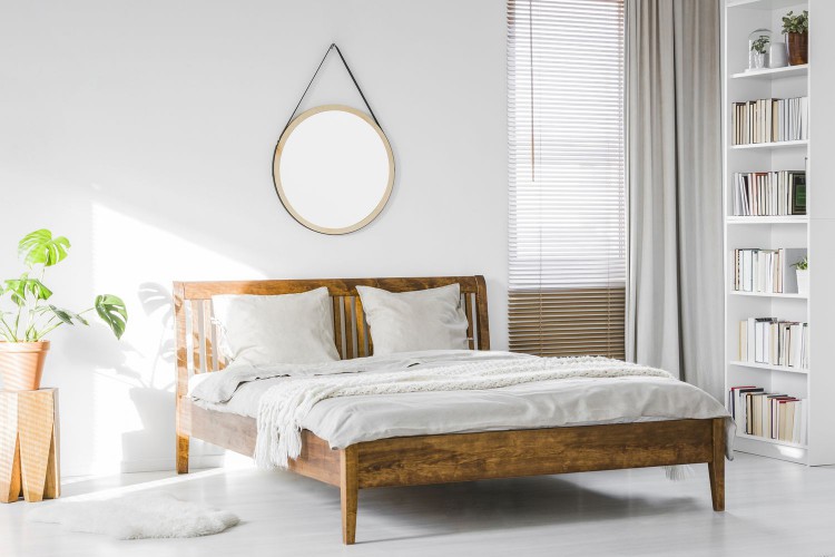 A wooden rustic bed frame and a home library bookcase in a natural, sunlit hotel room interior with white walls