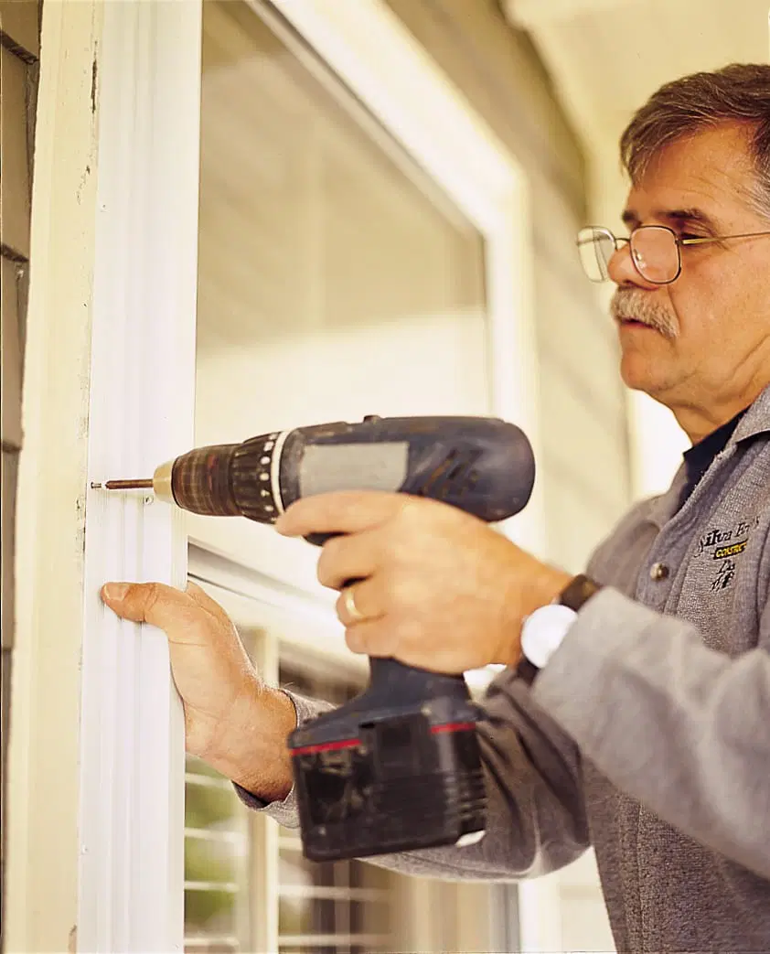 Image of Tom Silva installing storm windows on a home, driving screws into the perimeter of the storm window to secure it. 