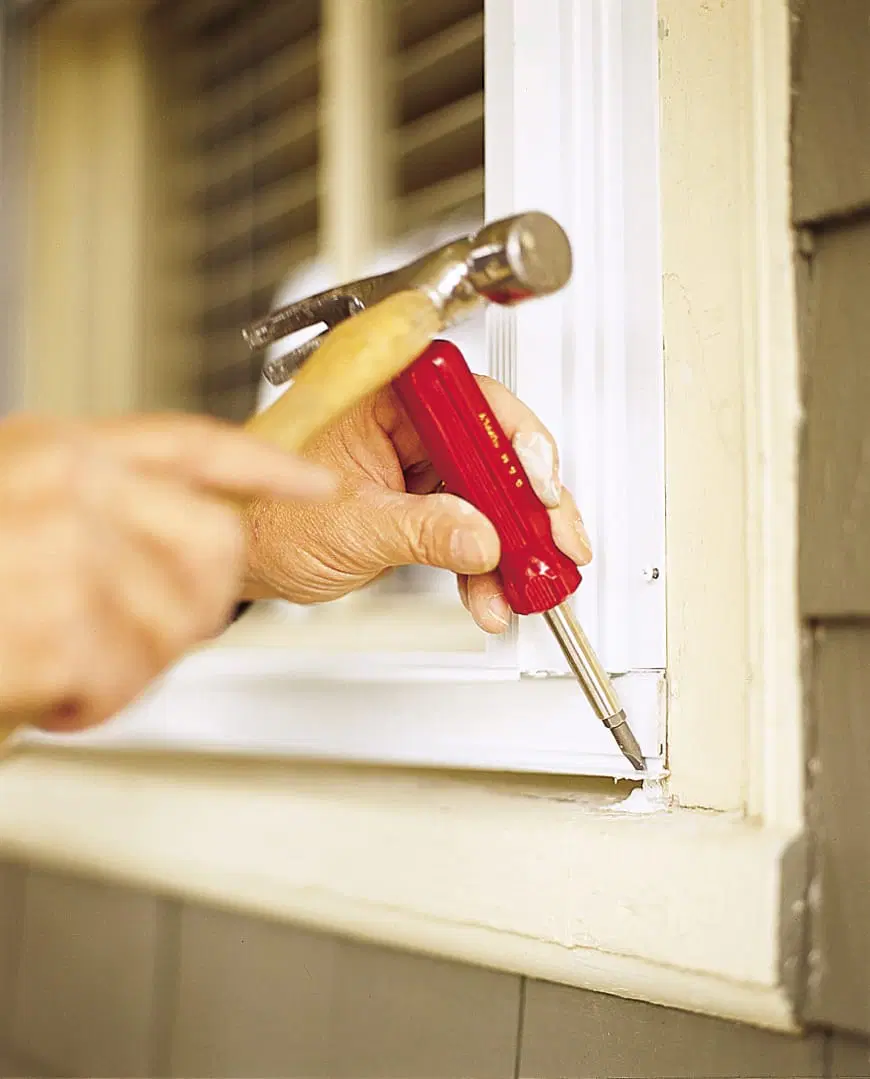 Image of Tom Silva installing storm windows on a home, using a hammer to tap the bottom sill extender until it rests against the sill. 
