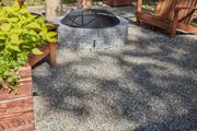 Pea gravel surrounding circular gray-stoned fire pit with wooden furniture and plants nearby