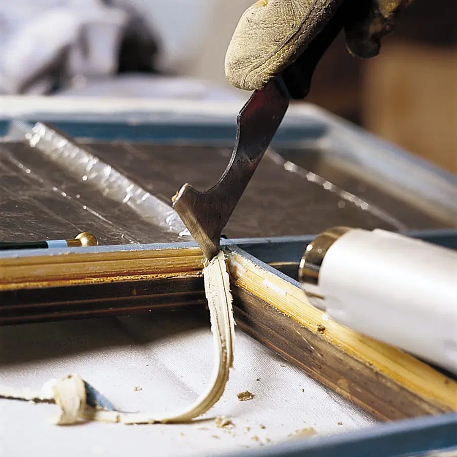A person using a painter tool to remove loose putty from a window pane.