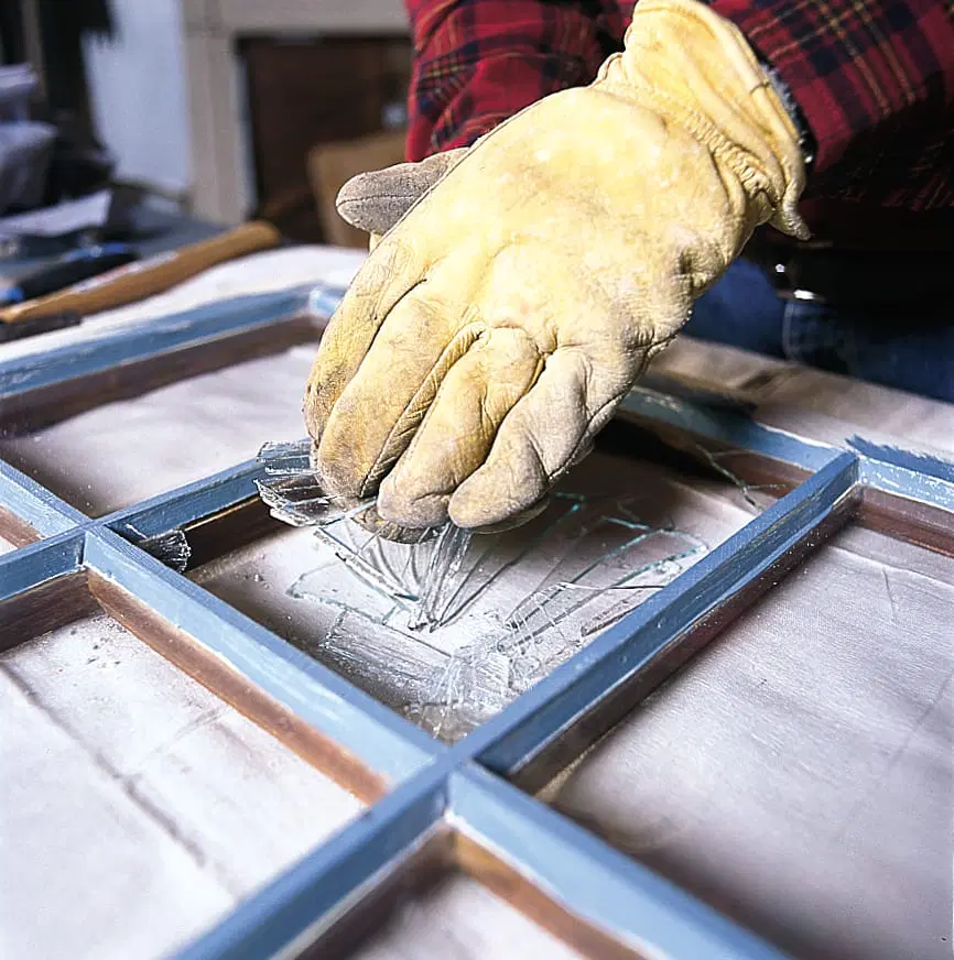 A person wearing a glove to remove broken glass from a window pane.