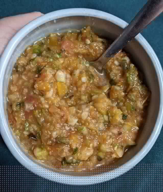 An image of yellow/orange rough chopped soup with some recognizable cucumber and tomato in it, in a light blue ceramic bowl.