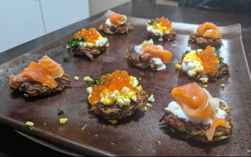Latkes on a board with smoked salmon, hard boiled egg, row, chives, and creme fraiche. 