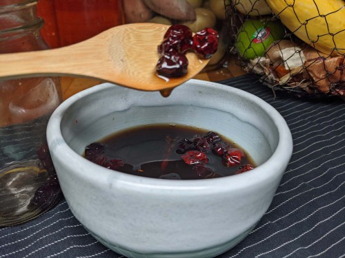 A wooden spoon dripping red honey and cranberries over a bowl of fermented cranberries. 