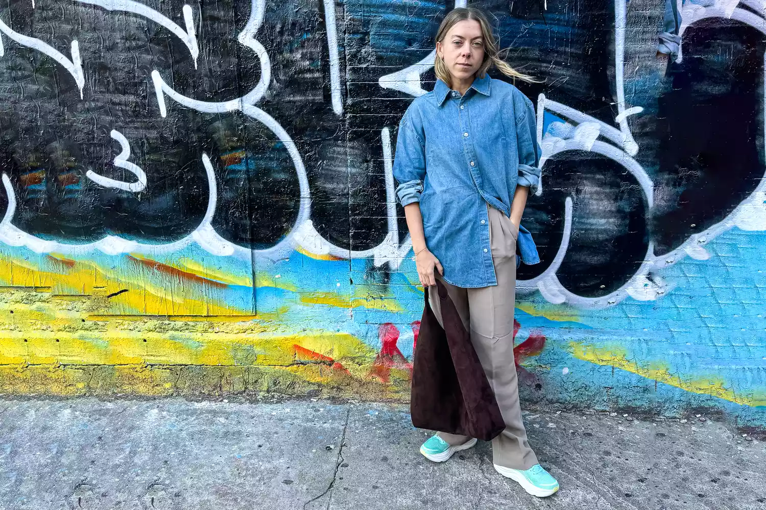 A woman stands against a wall wearing a denim shirt, pants, and Hoka Bondi 9 sneakers