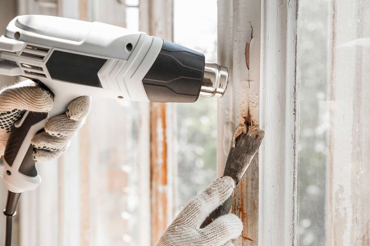 person removing old paint from a window with a heat gun and scraper