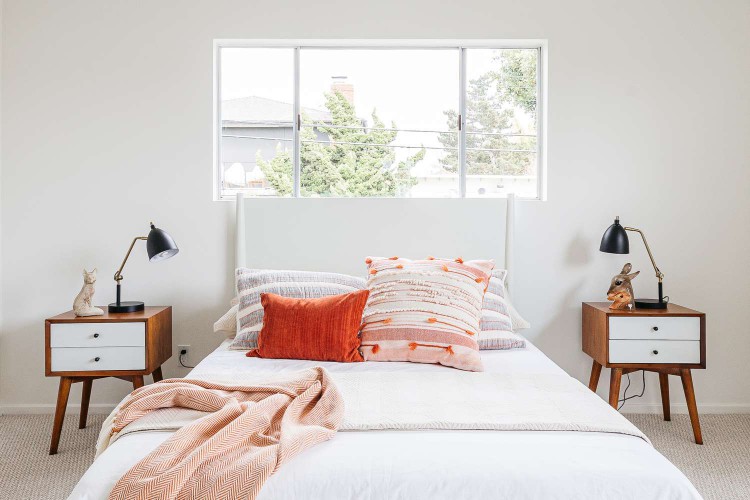 Minimalist bedroom with white walls, white bed with red cushions and two wooden night stands with white shelves