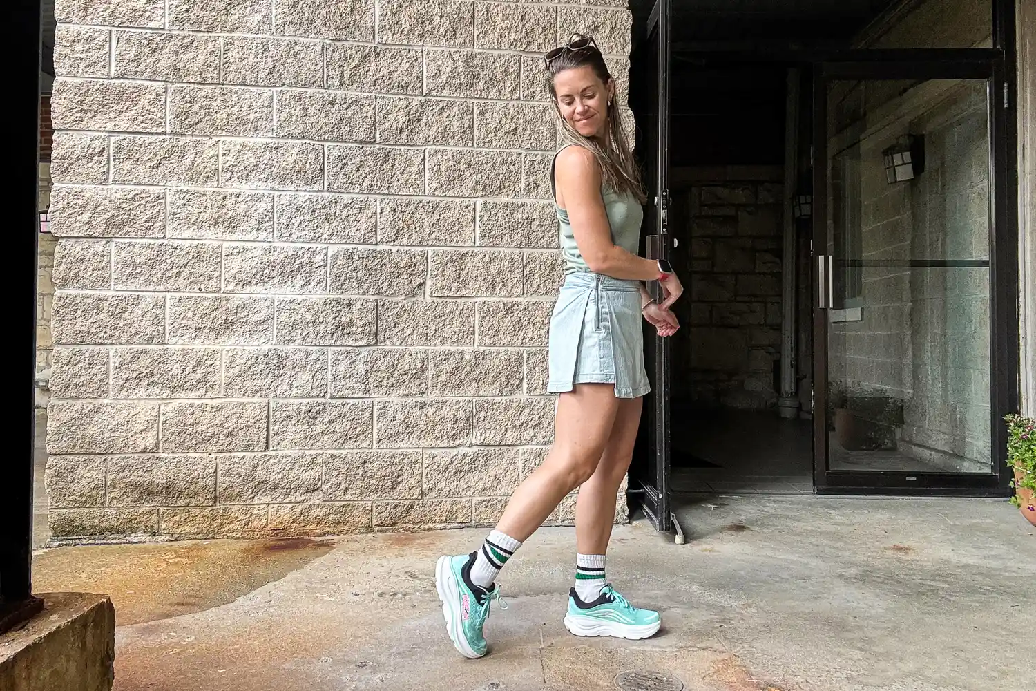 A person standing outdoors near a brick wall and a glass door wearing casual athletic attire and teal sneakers