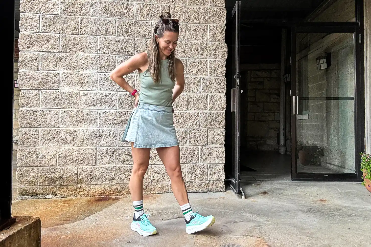 A person standing outdoors near a brick wall next to an open glass door smiling and looking down while wearing athletic attire and sneakers