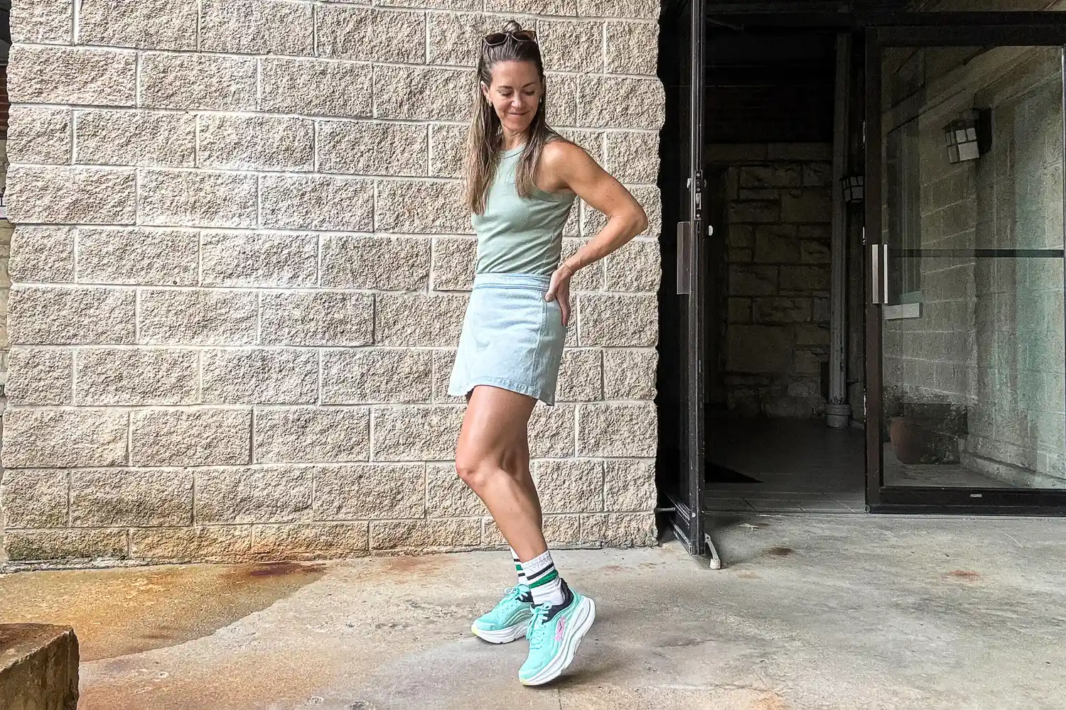 A woman standing outdoors next to a brick wall wearing casual clothing and sneakers with her hand on her hip