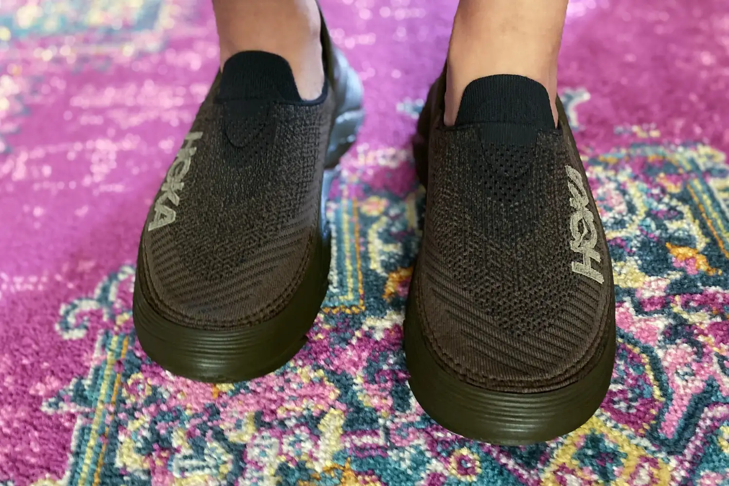 A close-up of two feet on a rug wearing Hoka Restore TC Recovery Shoes