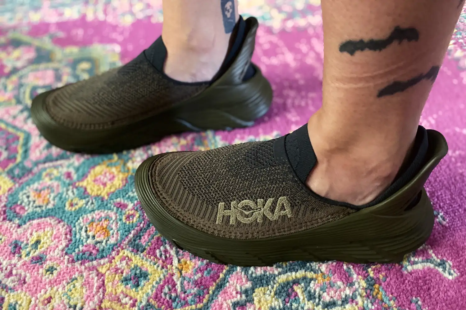 Two feet standing on a rug while wearing Hoka Restore TC Recovery Shoes