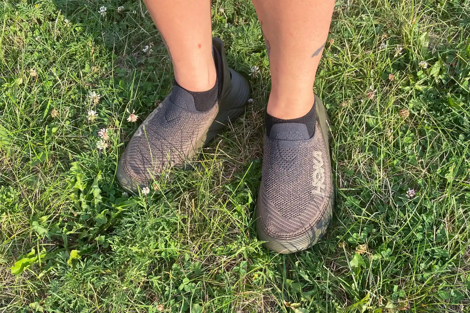 Two feet wearing Hoka Restore TC Recovery Shoes in grass