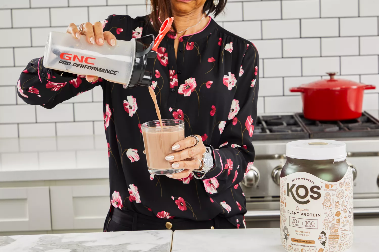 Hands pouring protein shake into a glass with KOS Vegan Superfood Protein Powder nearby on counter