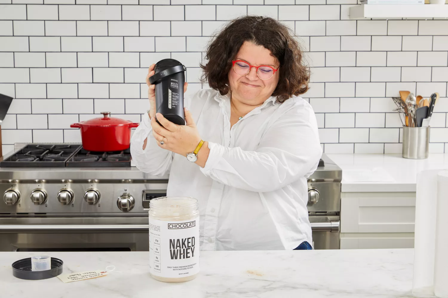 Person shaking a blender bottle next to a tub of Naked Whey Chocolate Protein Powder