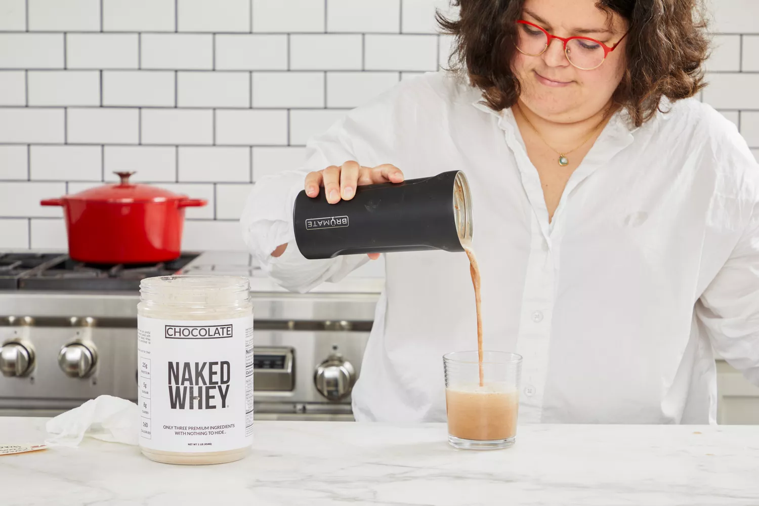 Person pouring a shake into a glass next to tub of Naked Whey Chocolate Protein Powder