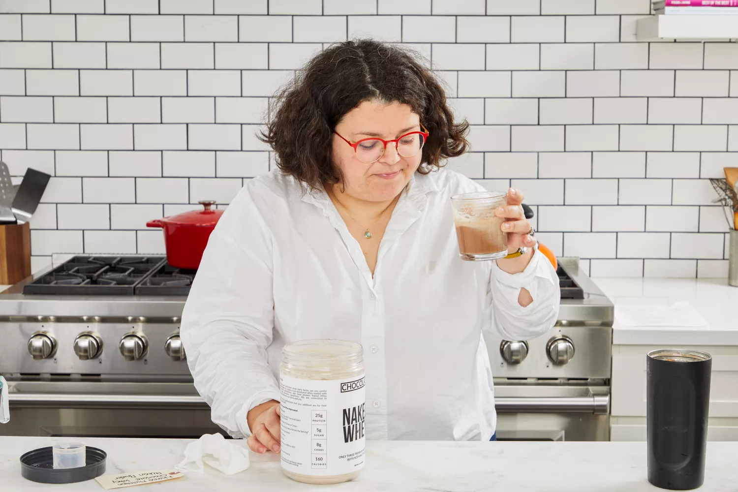 Person holding a protein shake with Naked Whey Chocolate Protein Powder container on counter
