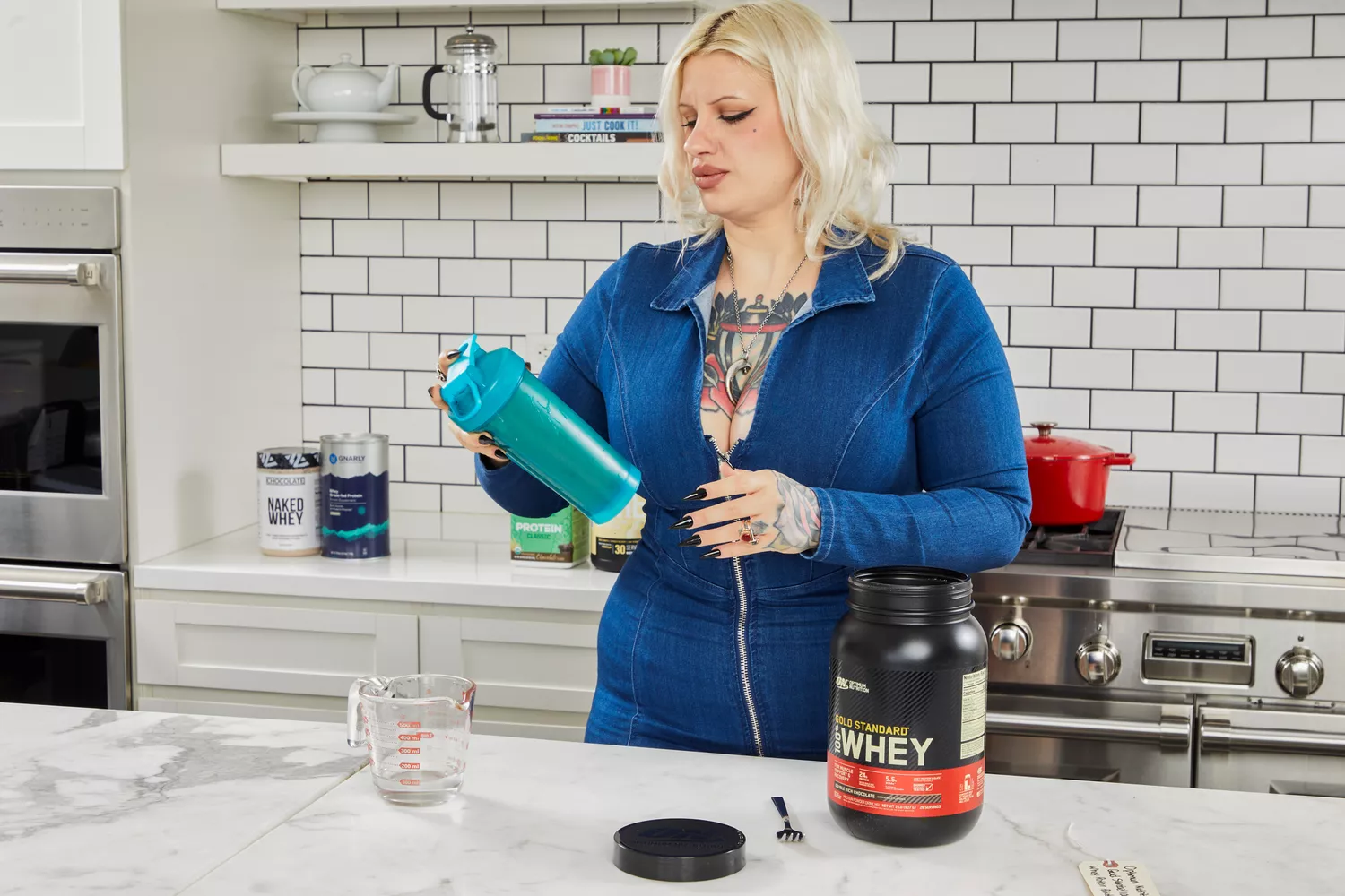 A person holding a shaker bottle with a tub of Optimum Nutrition Gold Standard 100% Whey Protein Powder on counter nearby