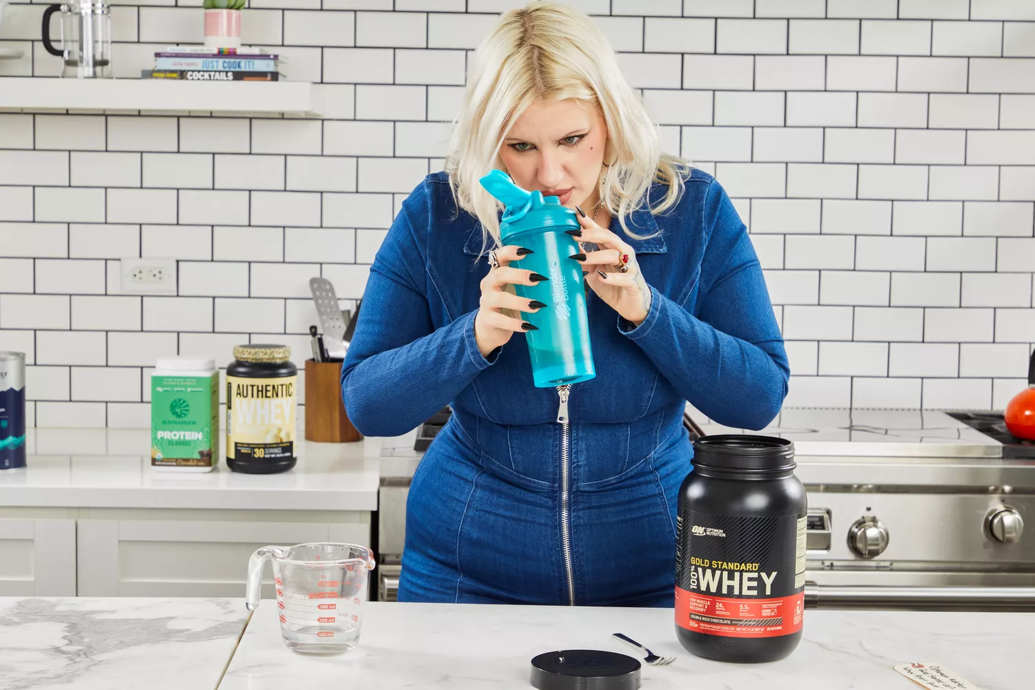 A person sniffing a shaker bottle with Optimum Nutrition Gold Standard 100% Whey Protein Powder nearby on counter