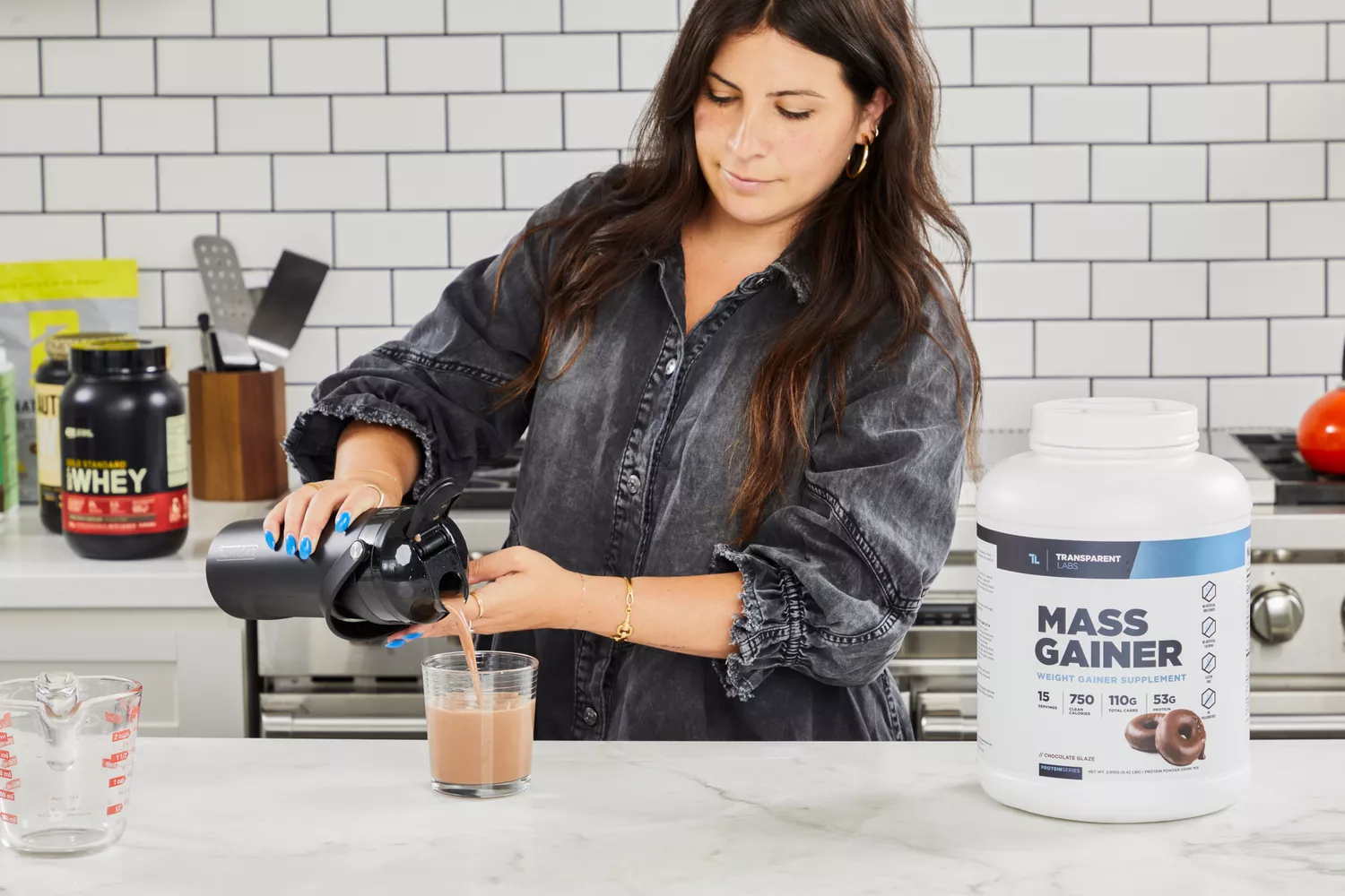 A person pouring a protein shake into a glass next to tub of Transparent Labs Mass Gainer Weight Gainer Supplement