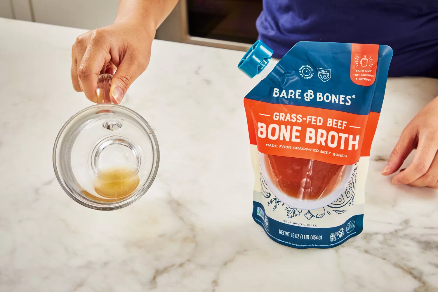 hand holds glass of Bare Bones Grass-Fed Beef Organic Bone Broth with pouch on countertop