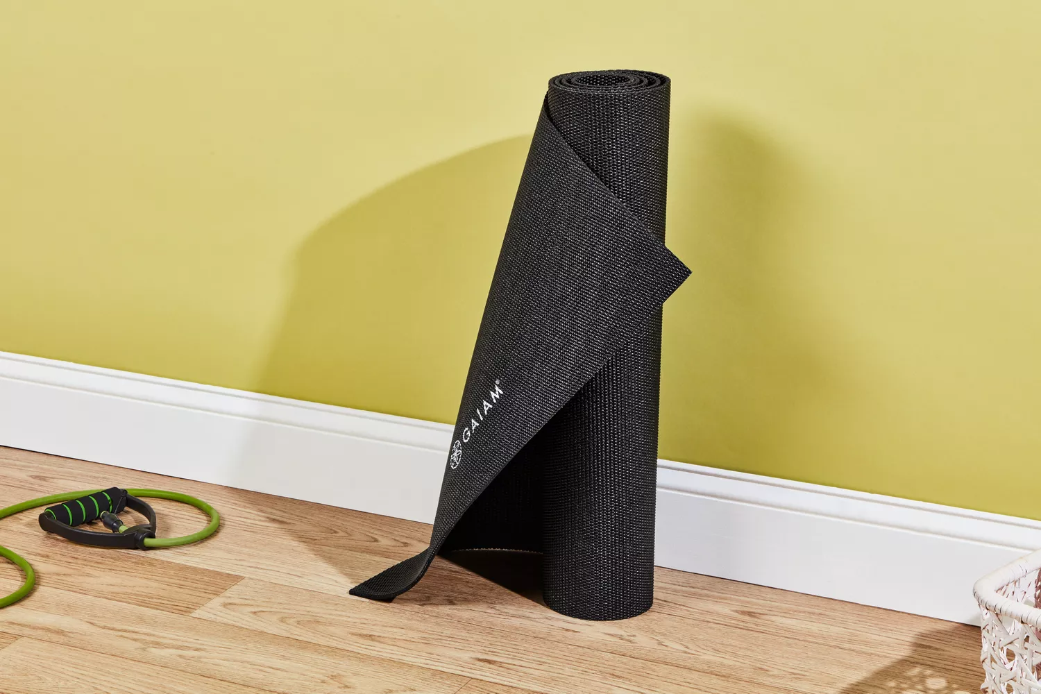 A rolled up Gaiam Classic Solid 5mm Yoga Mat lies vertically against a yellow wall