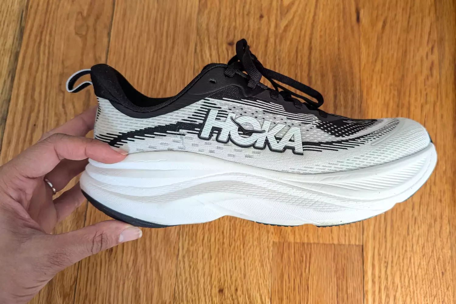 A hand holding a Hoka Women's Skyflow sneaker over a wooden surface