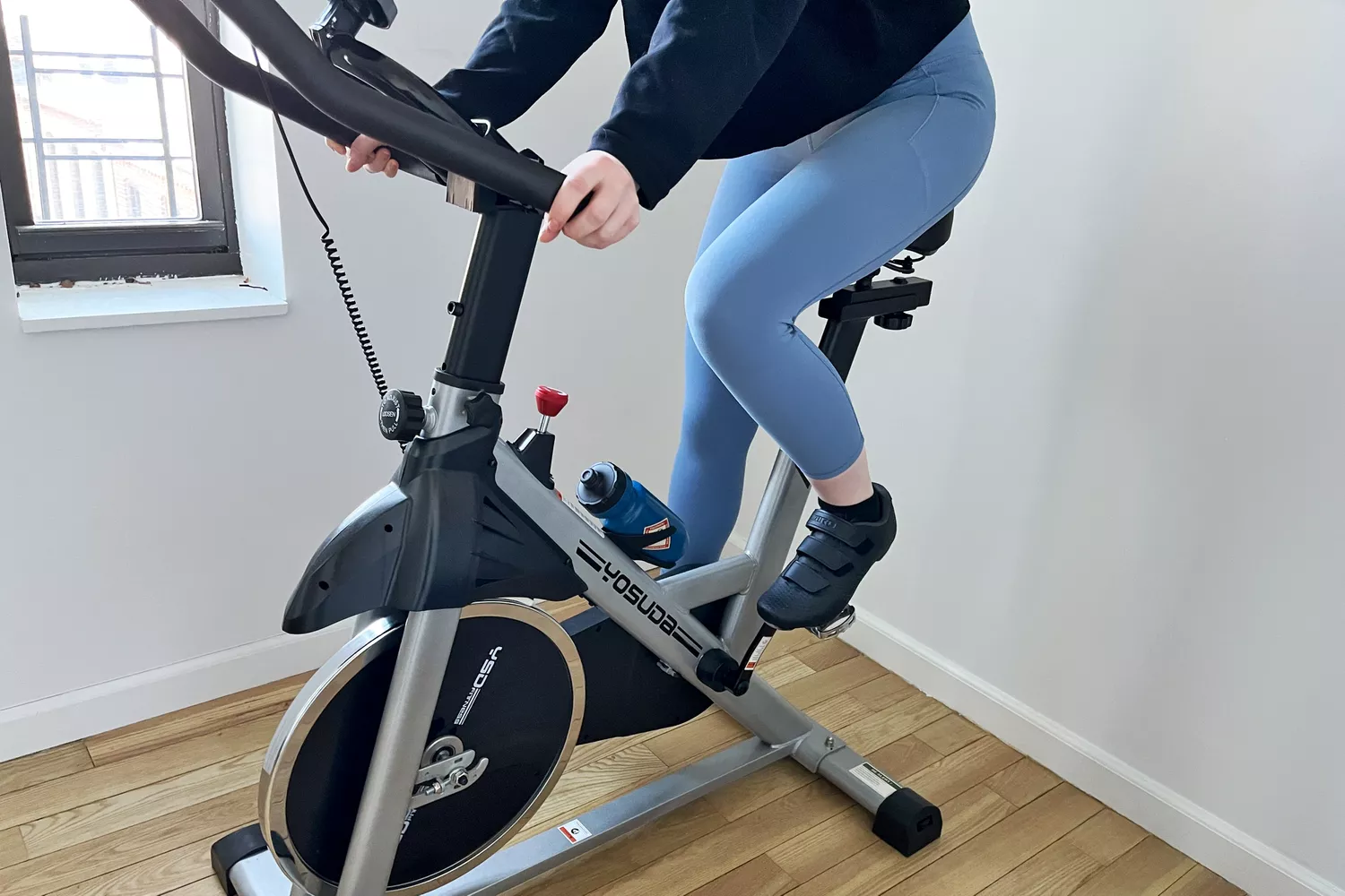 A person exercising on YOSUDA Indoor Cycling Bike in a white room with hardwood floors