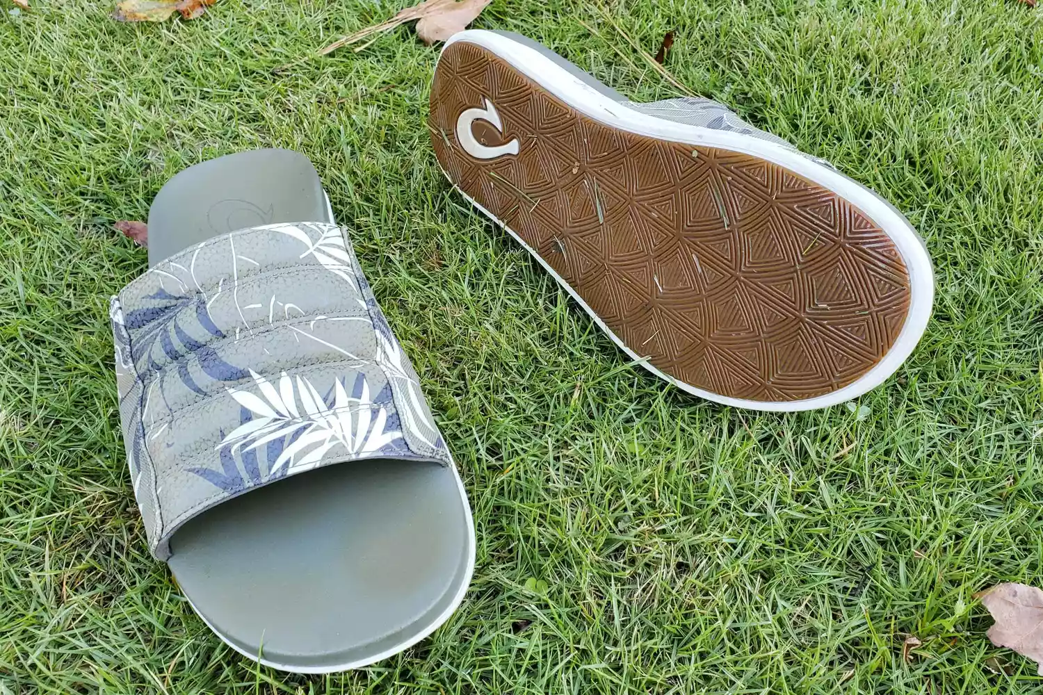 A pair of OluKai Maha’Olu slides on grass with one sole displayed