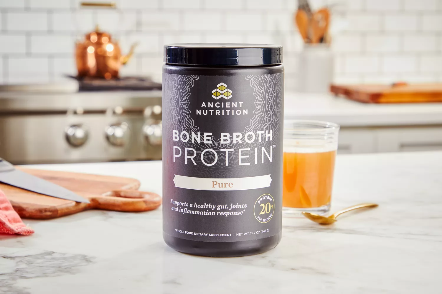 A canister of Ancient Nutrition Bone Broth Protein with glass of broth on countertop