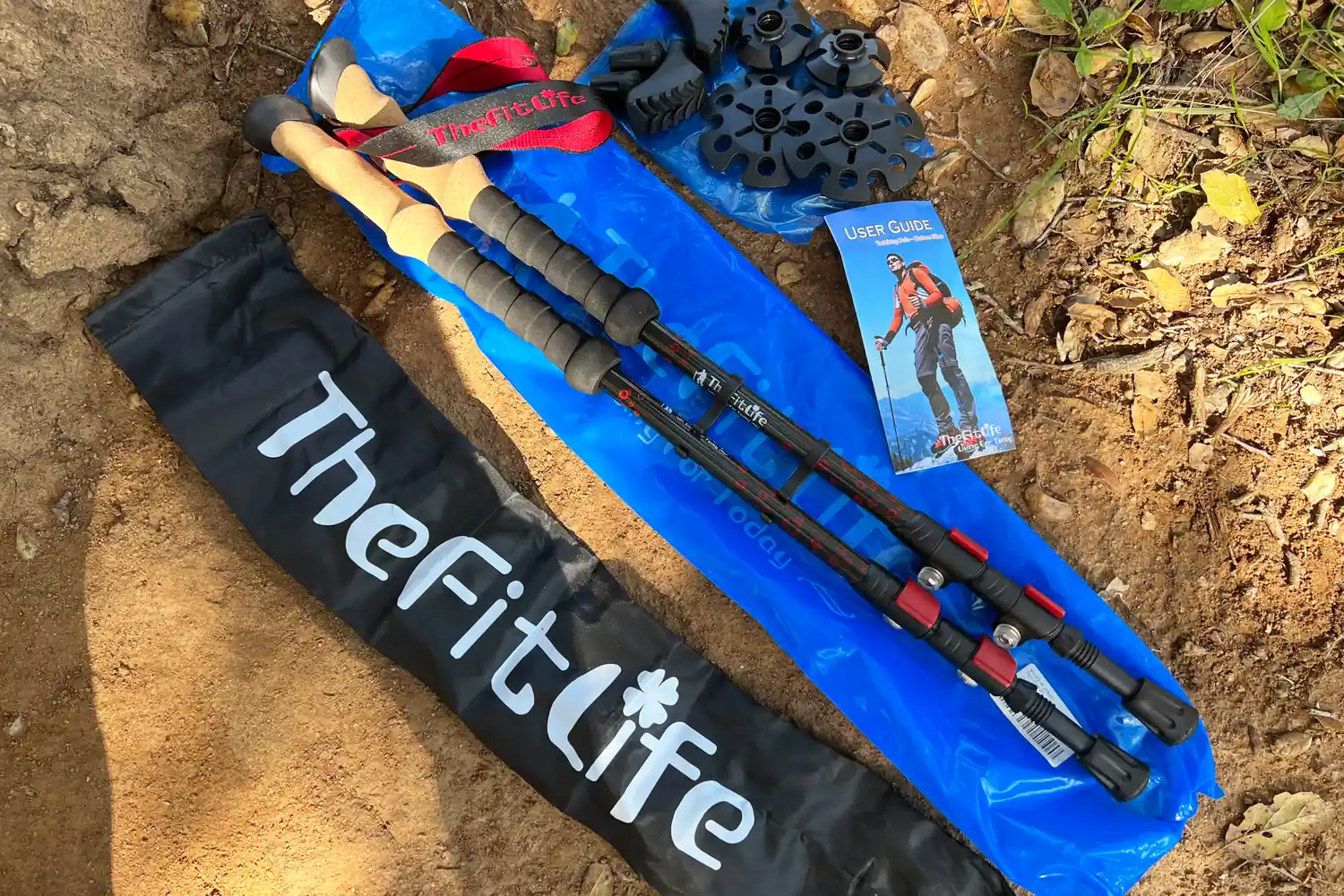 Set of trekking poles with accessories and a carrying bag labeled TheFitLife and instructional manual on a dirt background