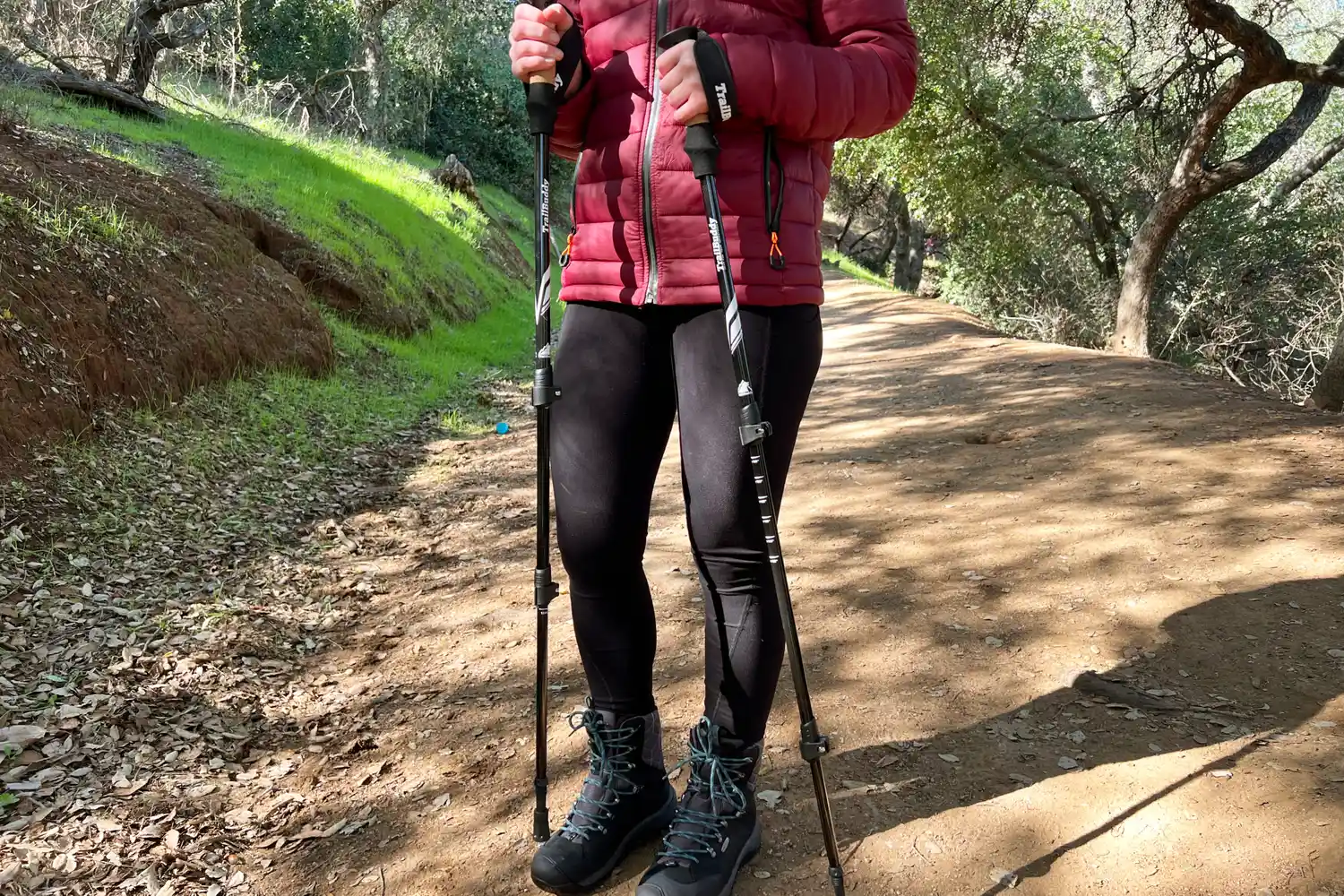 A person standing on a trail holding hiking poles with visible hiking boots and outdoor gear