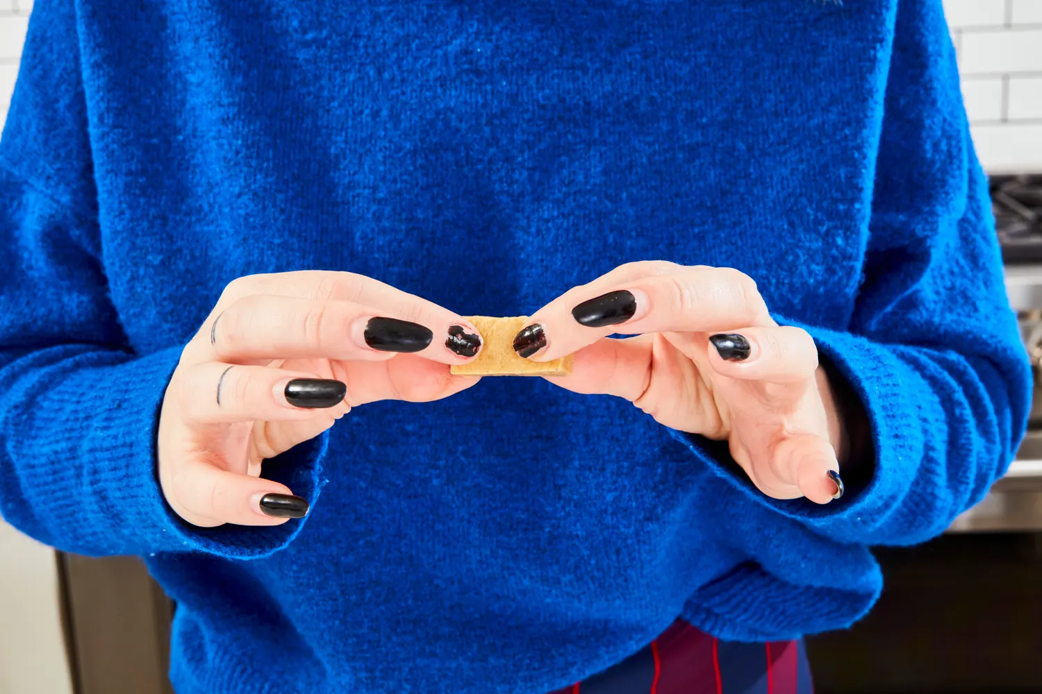 A person holding a small rectangular piece of a food item between their hands with black painted nails background includes a kitchen scene