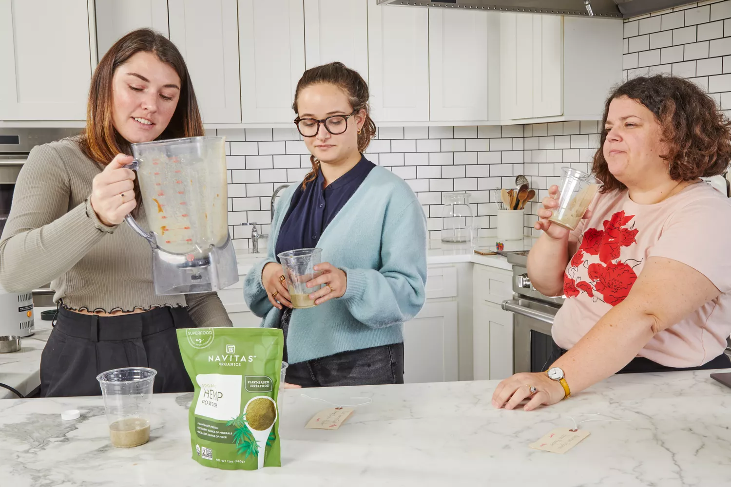 Three women holding plastic cups in front of Navitas Organics Hemp Powder on marble counter