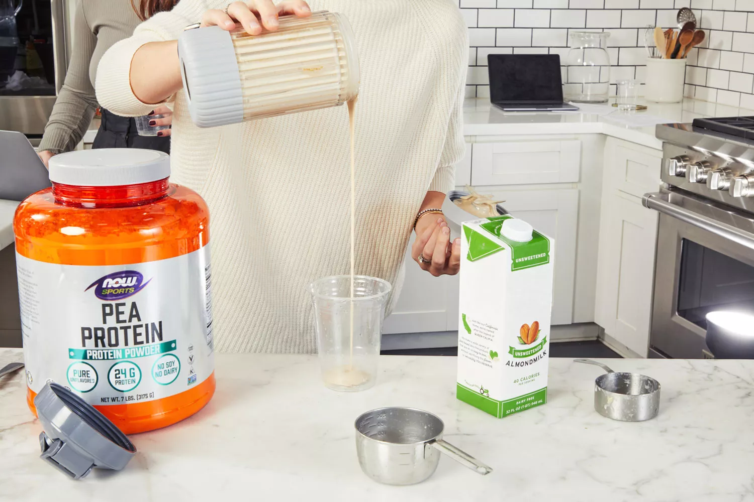 Hands pouring smoothie into a plastic cup next to NOW Sports Pea Protein and almond milk