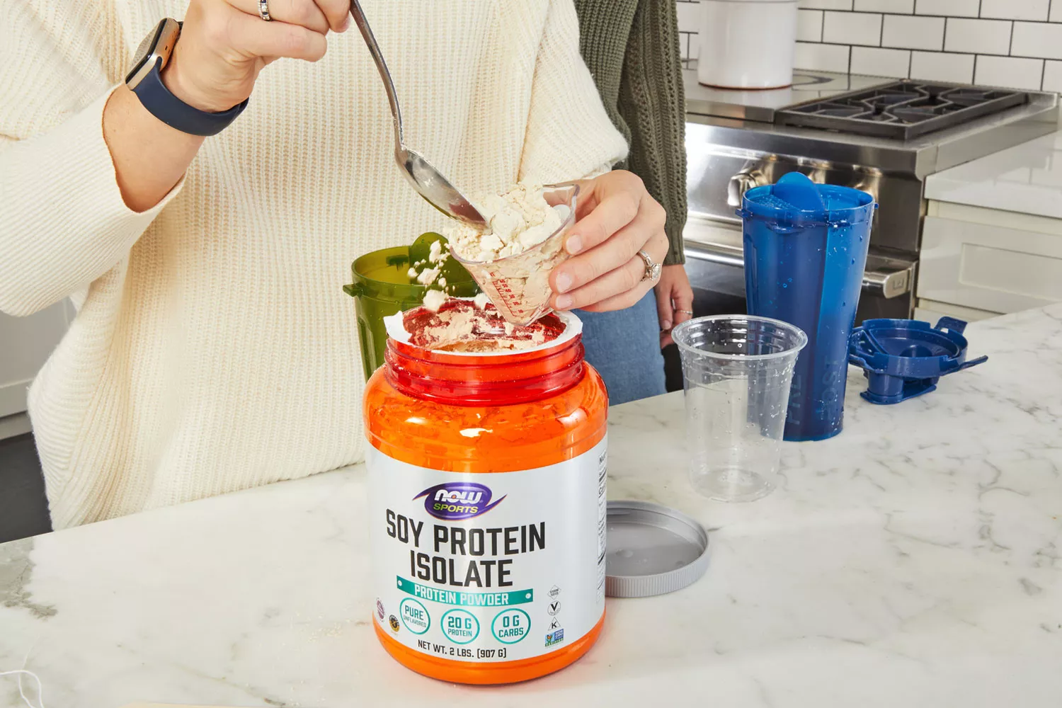 Hands measuring out NOW Sports Pea Protein from package with blender bottle in background