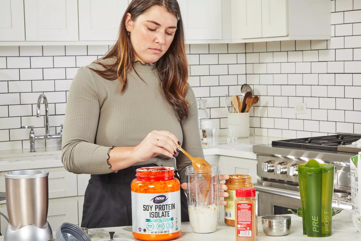 Person spooning peanut butter into blender bottle next to NOW Sports Pea Protein