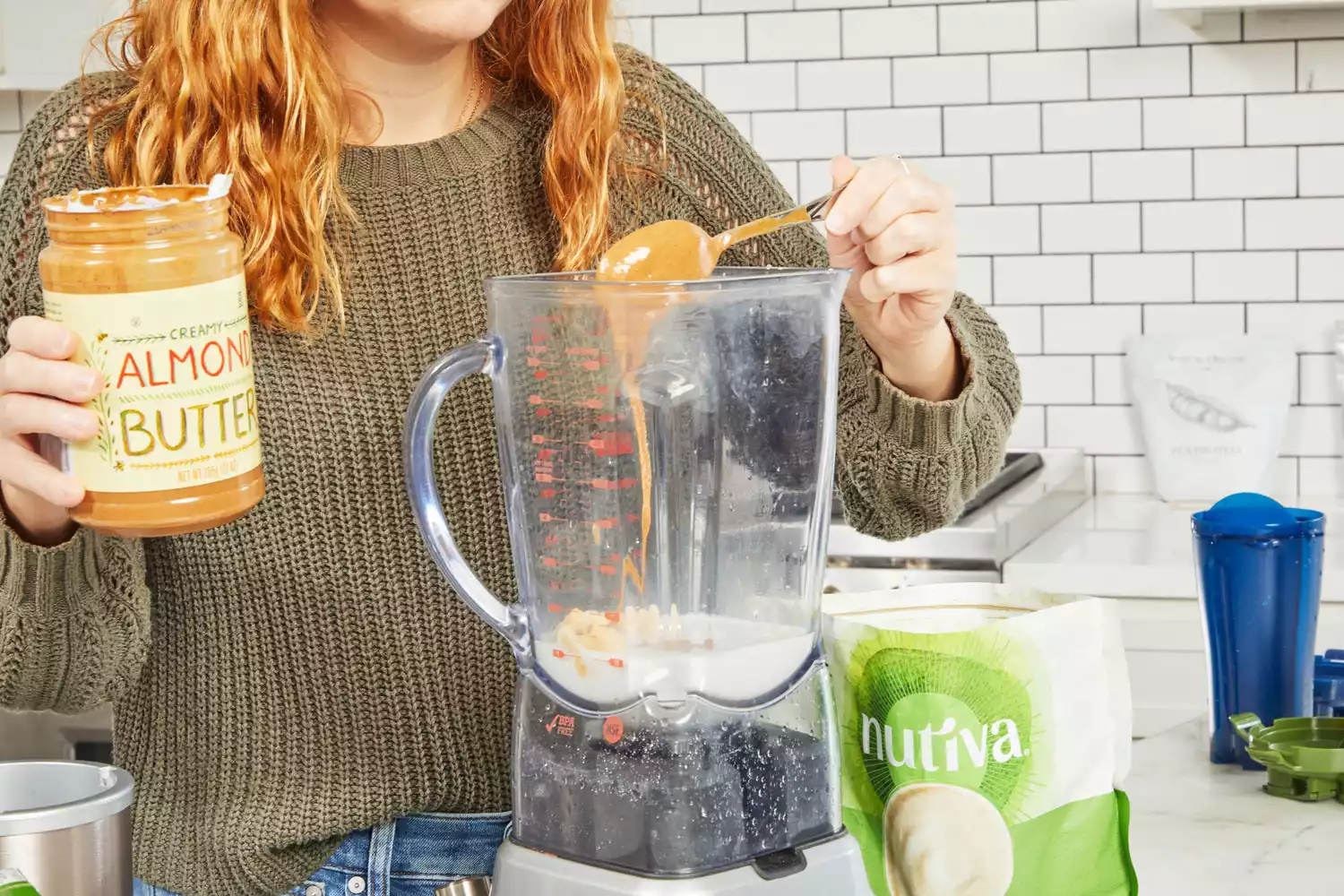 A hand spooning almond butter into a blender next to Nutiva Hemp Seed Protein Powder