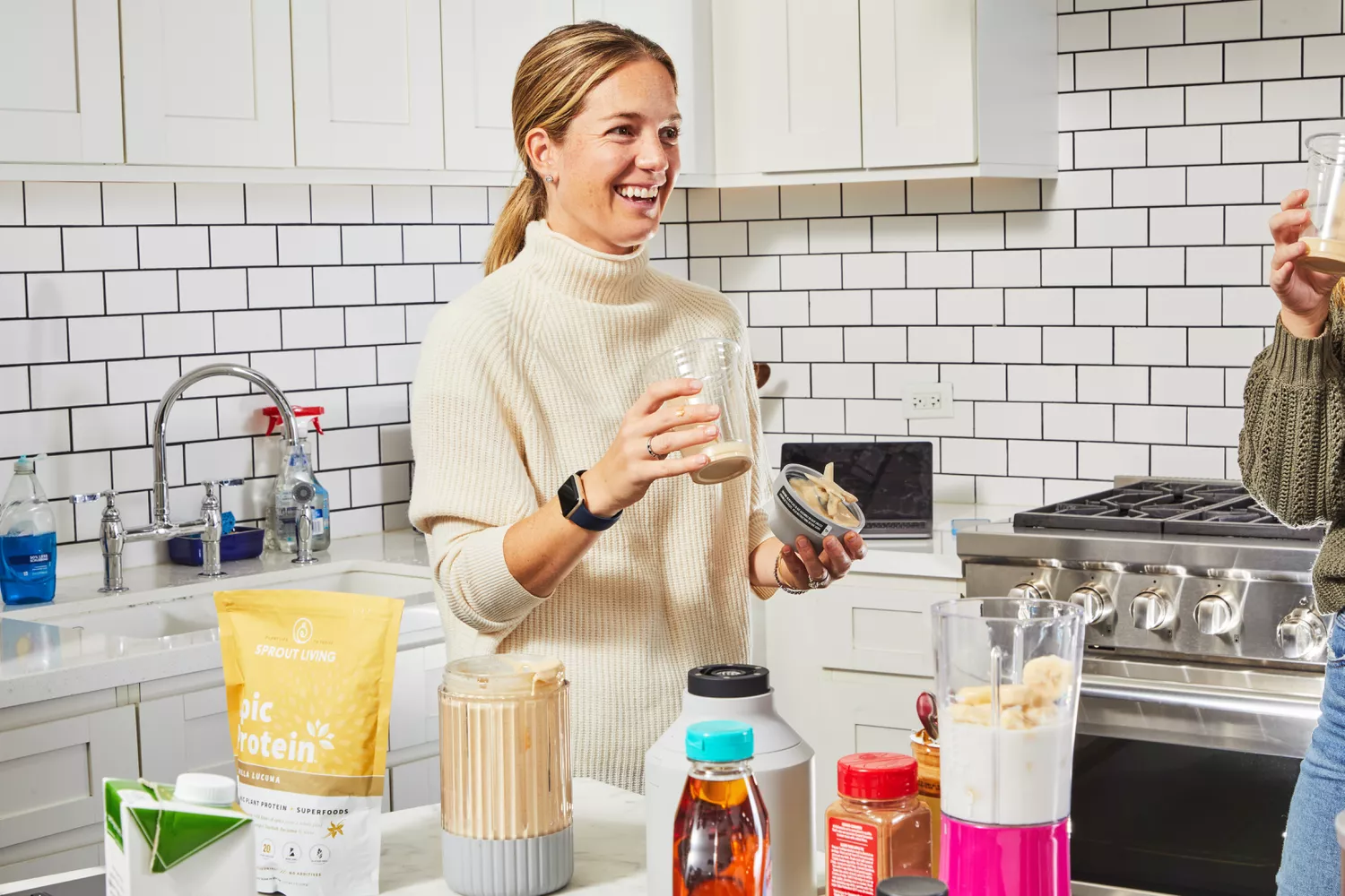 Person holding glass with Sprout Living's Epic Protein Vanilla Lucuma Powder, blender and various ingredients on counter