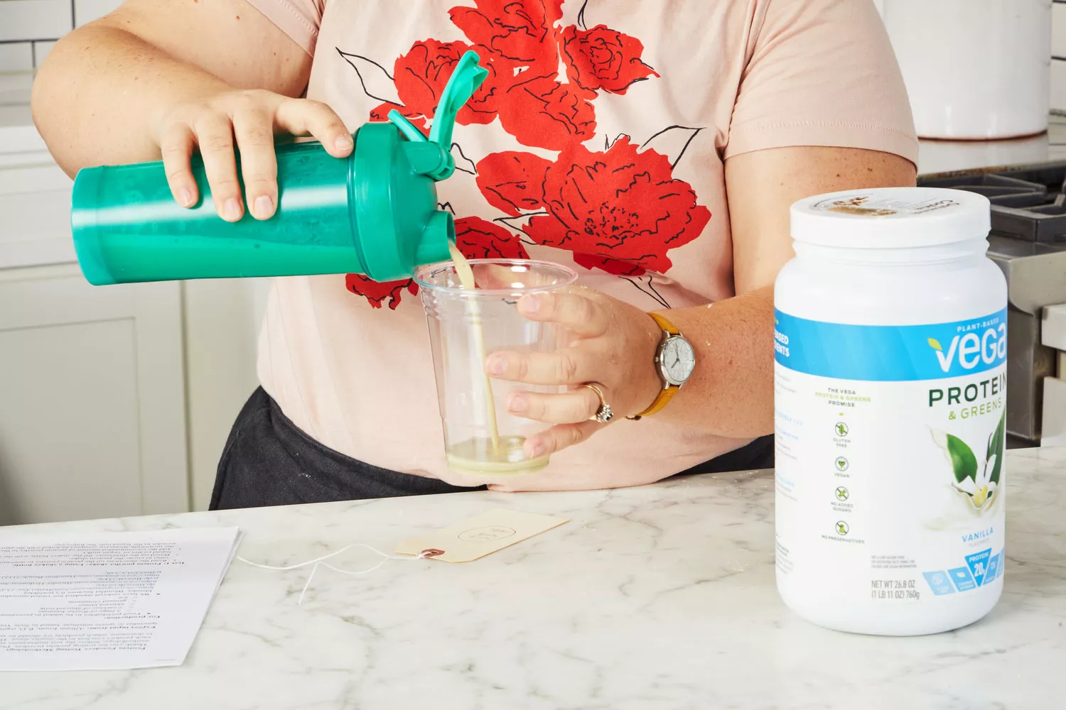 Hands pouring liquid from a protein shaker into a plastic cup next to Vega Protein & Greens Plant-Based Protein Powder