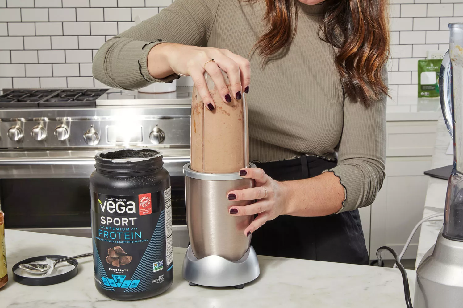 A hand on a blender next to container of Vega Sport Premium Protein on counter