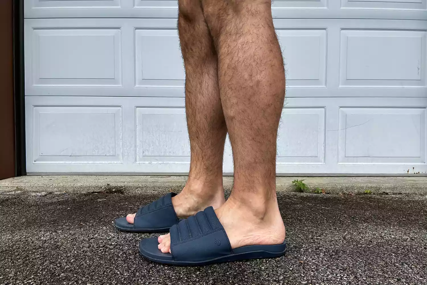 A person wears the OluKai MahaâOlu Menâs Slide Sandals outside on asphalt