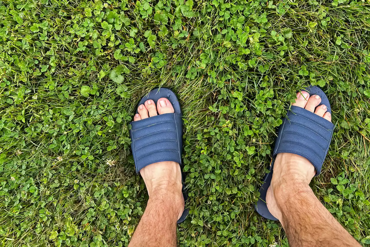 A person stands in grass while wearing the OluKai MahaâOlu Menâs Slide Sandals