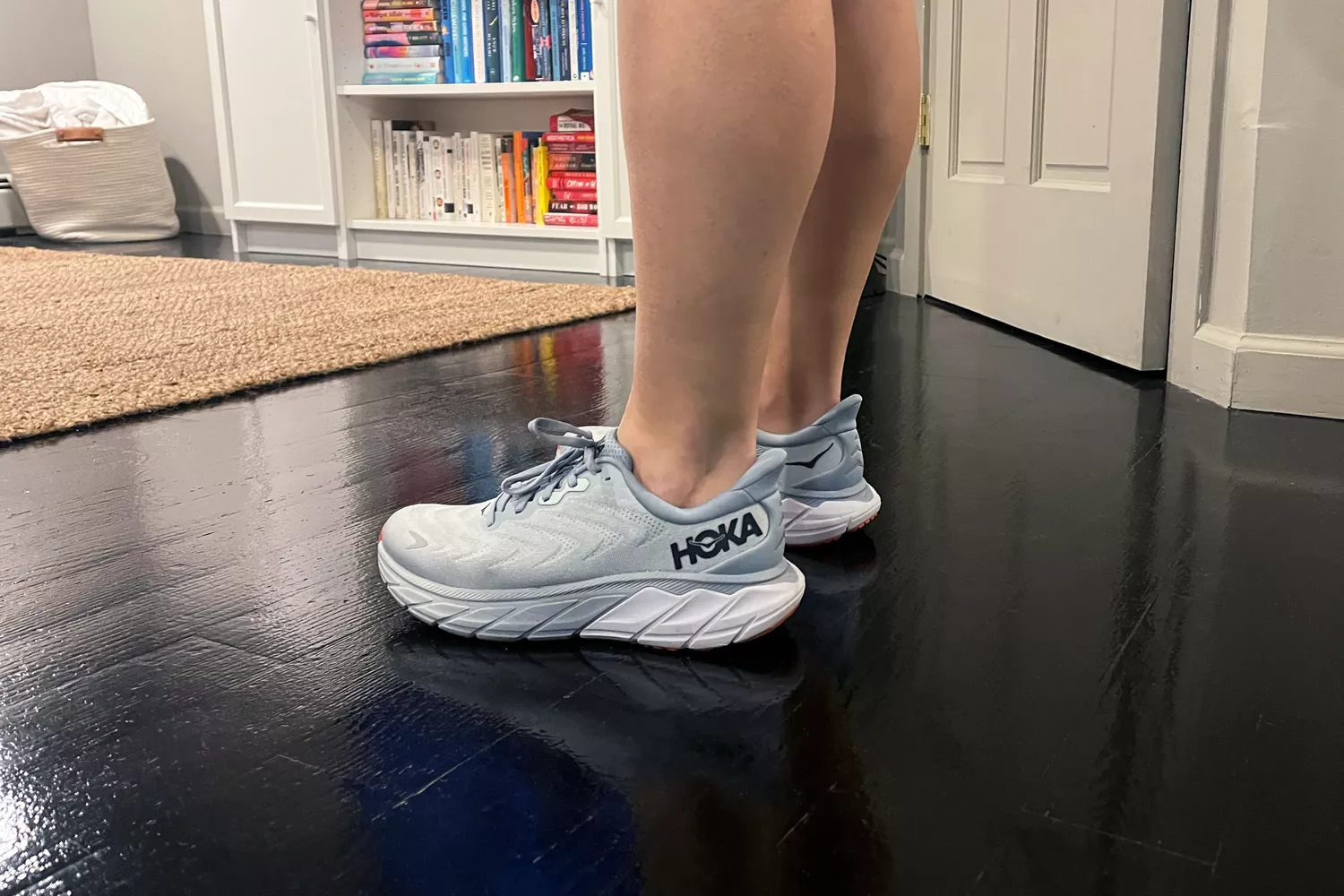Feet wearing the Hoka Women's Arahi 6 Running Shoe while standing on a wooden floor
