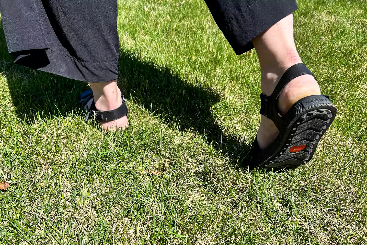A close-up of feet walking on grass while wearing the Sorel ONA AVE Women's Ankle Strap Flat Sandal