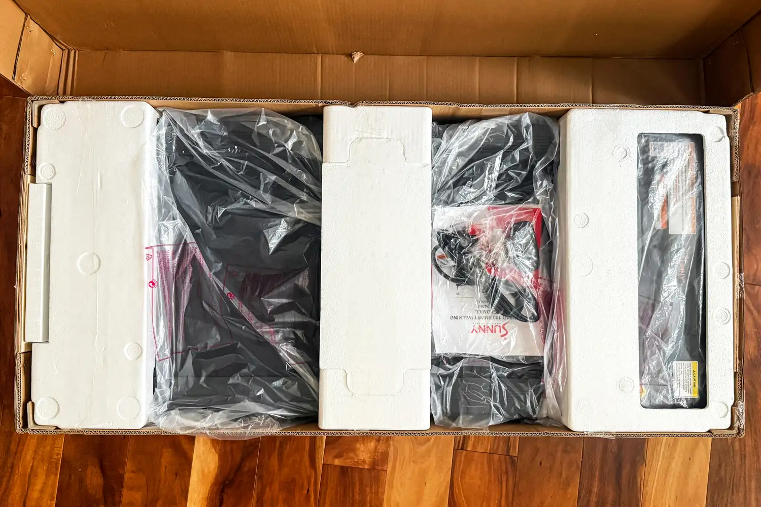 The Sunny Health & Fitness Treadpad 100 Smart Wi-Fi Connected Treadmill inside plastic packaging inside a cardboard box